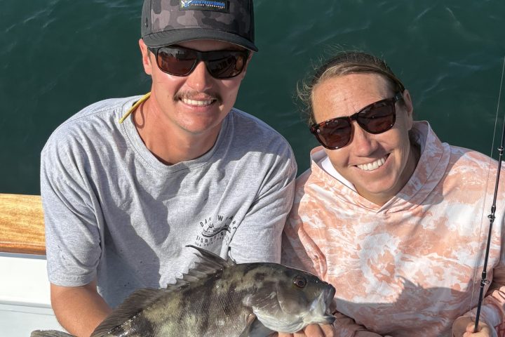 Two people on a boat holding a fish with fishing rods, smiling.