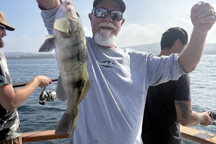 Man on boat holding a fish, smiling, with ocean in background.