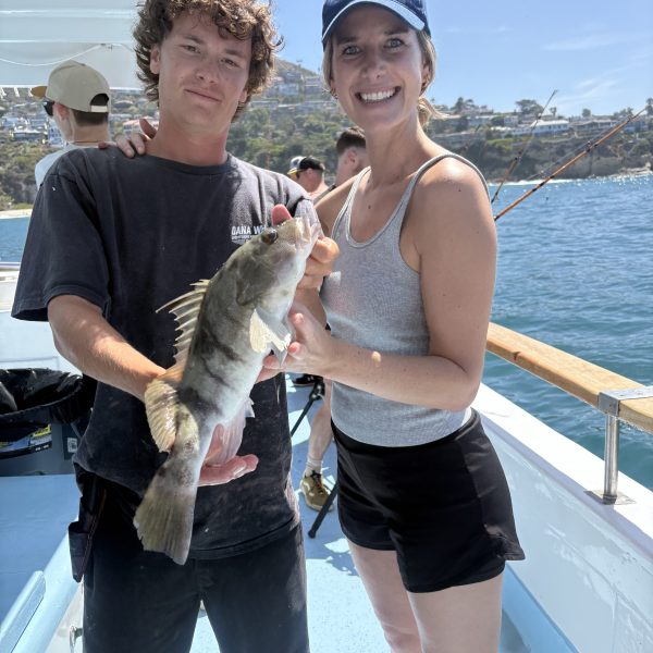 Two people on a boat holding a fish with a scenic coastline in the background.
