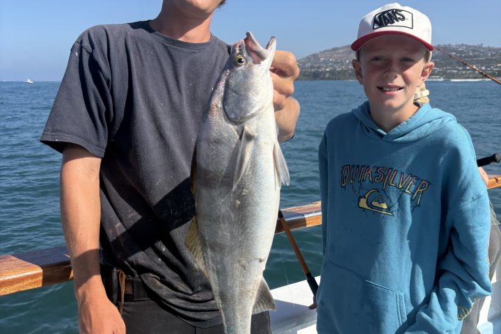 Two people on a boat holding a large fish, with a blue ocean background.