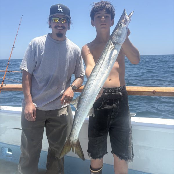 Two men on a boat holding a large barracuda fish.