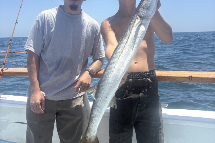 Two men on a boat holding a large barracuda fish.
