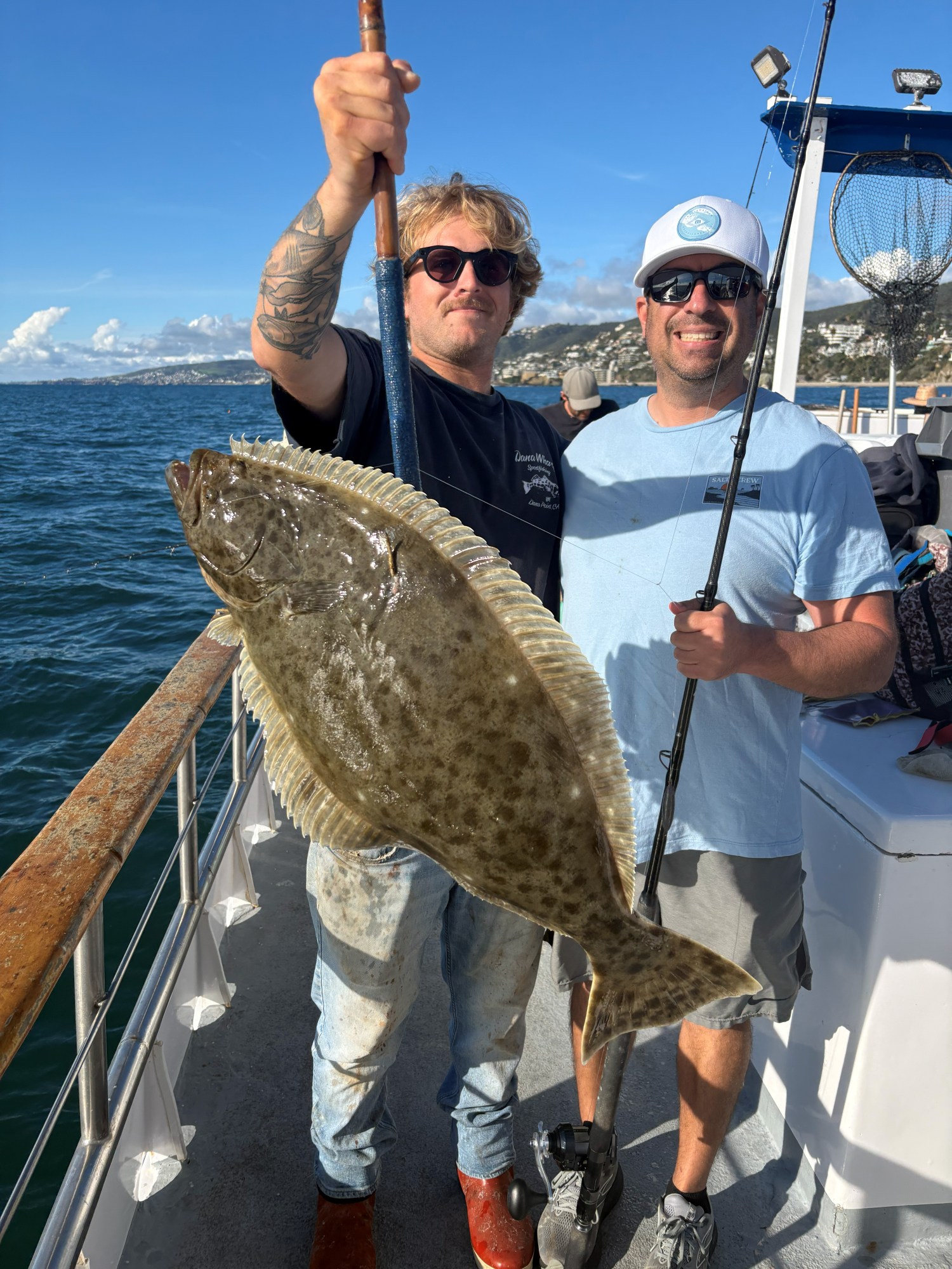 Two men on a boat holding a large fish with a blue sky and ocean in the background.