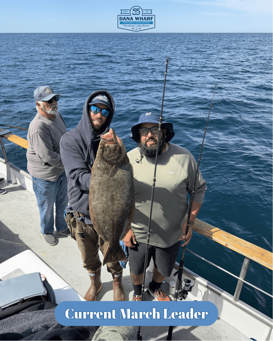 Three men on a boat holding a large fish with ocean in background.