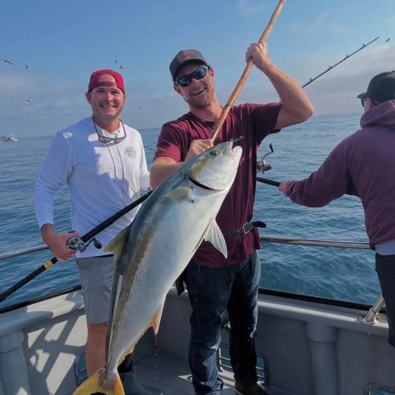 Two people on a boat holding a large fish they caught, with ocean and seagulls in the background.