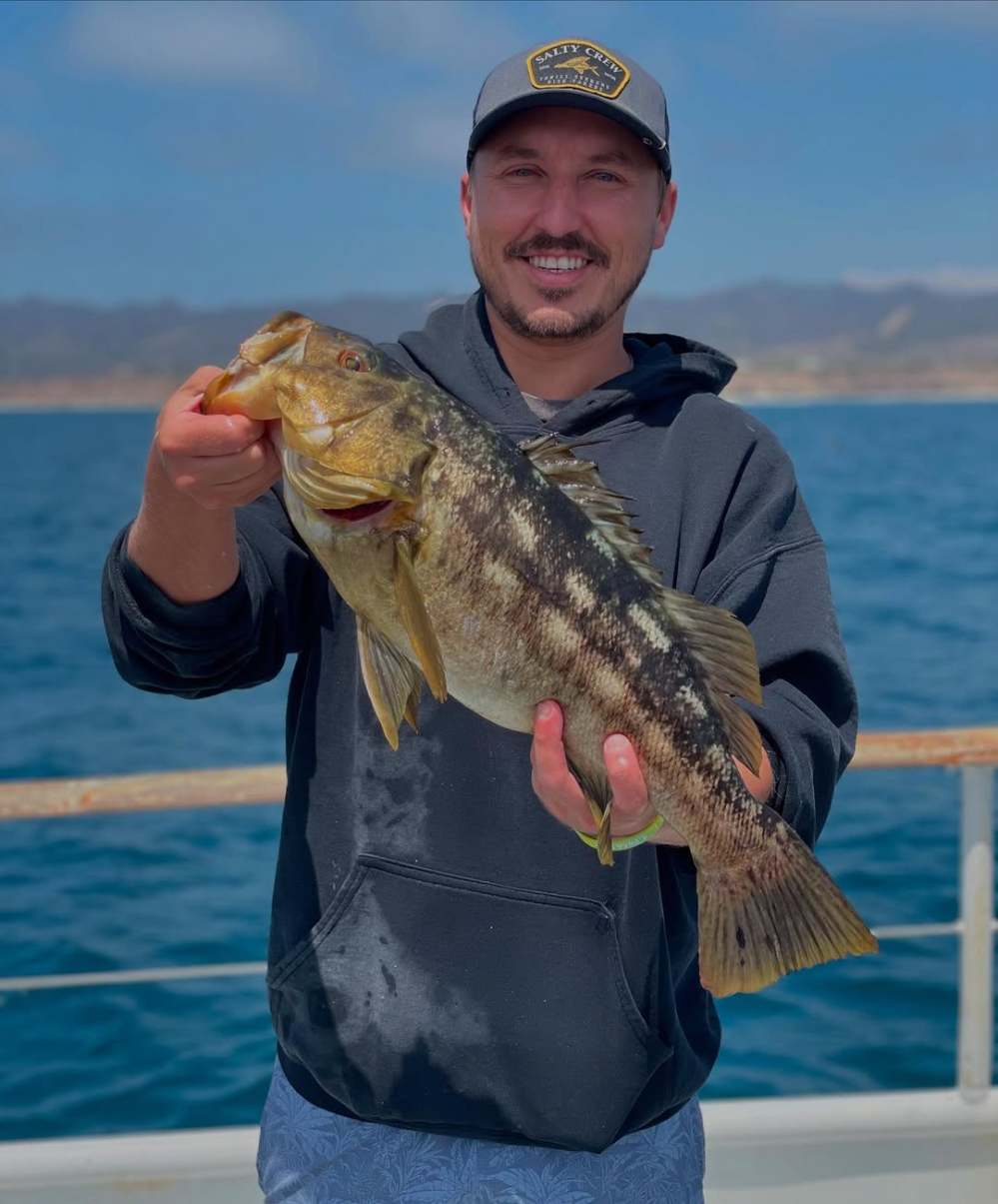 Man in cap holding a large fish, standing on a boat with ocean and mountains in background.