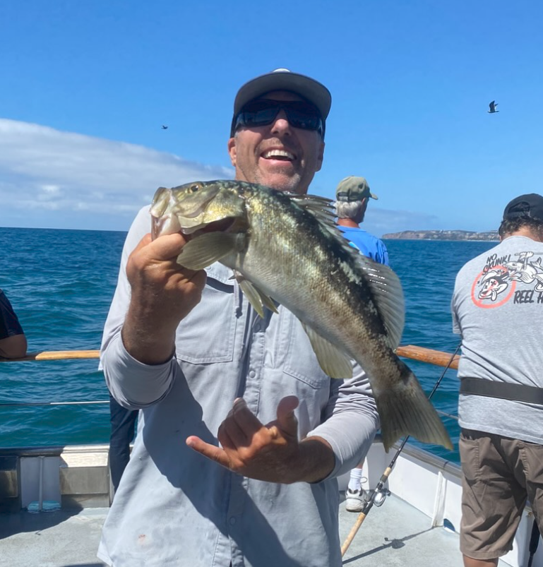 Man on boat holding fish, smiling, with ocean and clear sky in background.