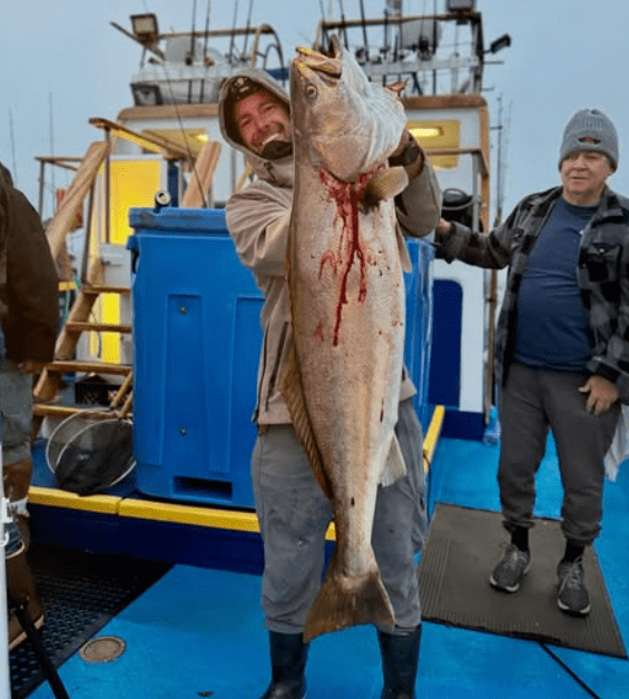 Person in hoodie holding a large fish on a boat deck. Another person stands nearby in a beanie.