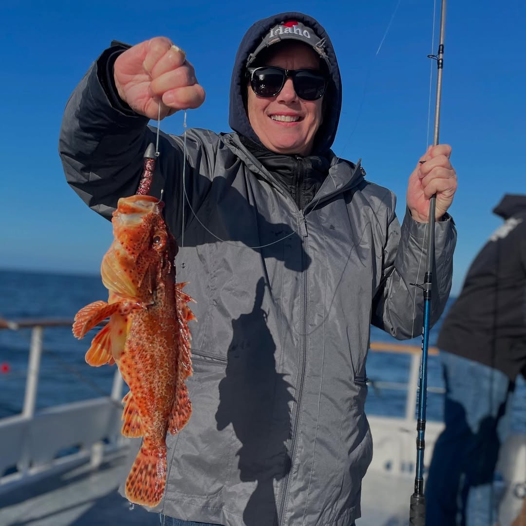 Person smiling, holding fishing rod and a fish on a boat with blue sky background.