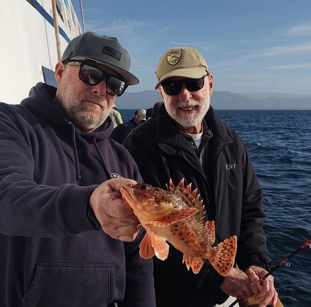 Two men on a boat, one holding an orange fish, with ocean and mountains in the background.