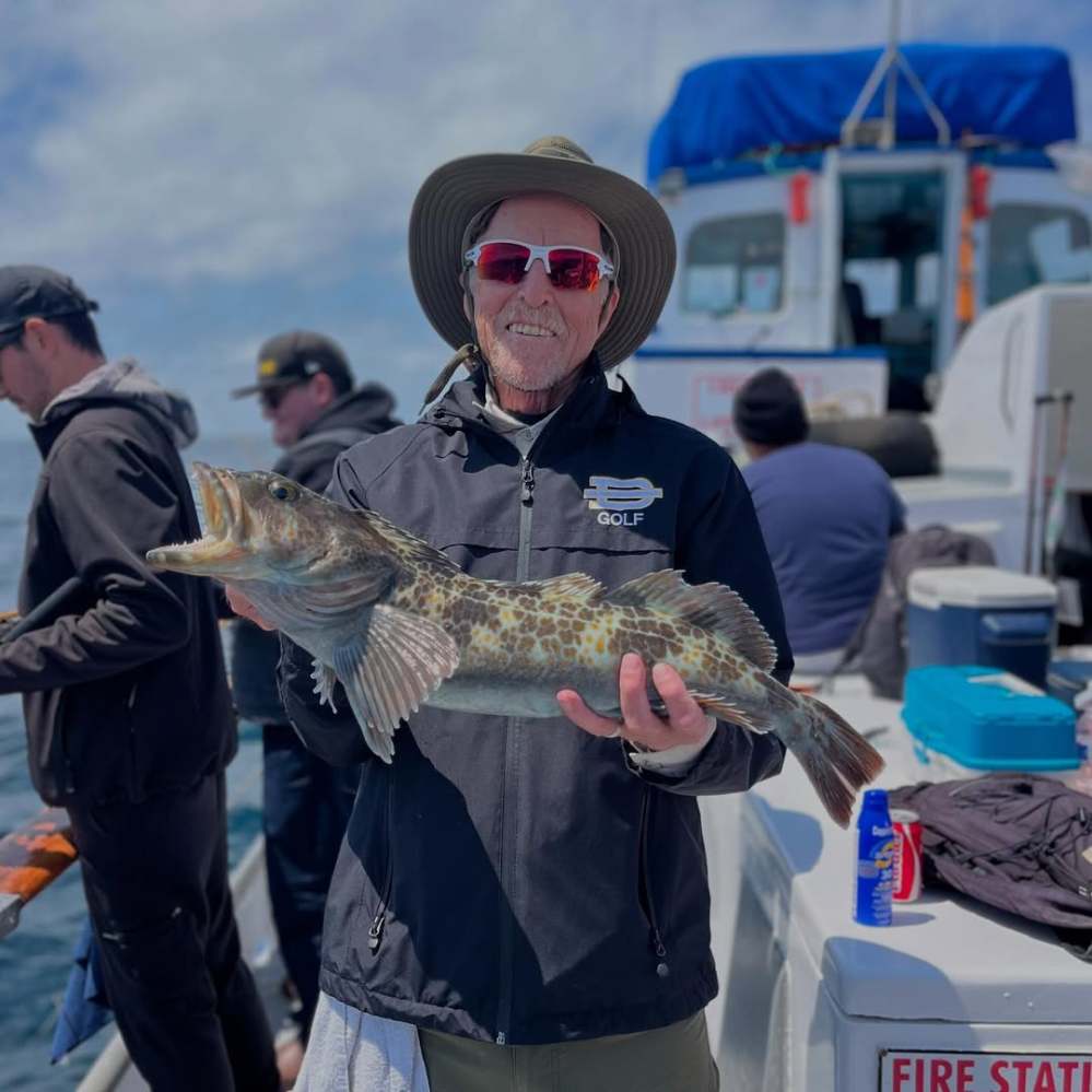 Man on boat holding large fish, wearing hat and sunglasses, with other people and equipment in background.