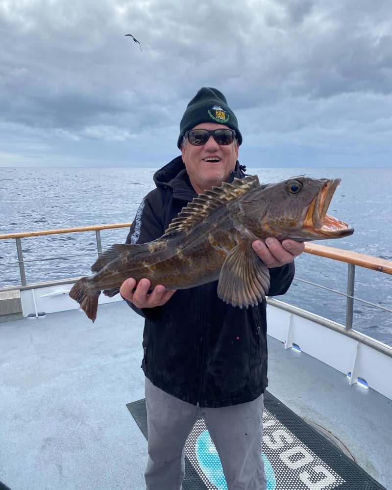 Person on boat holding a large fish with open mouth, cloudy sky and ocean in the background.
