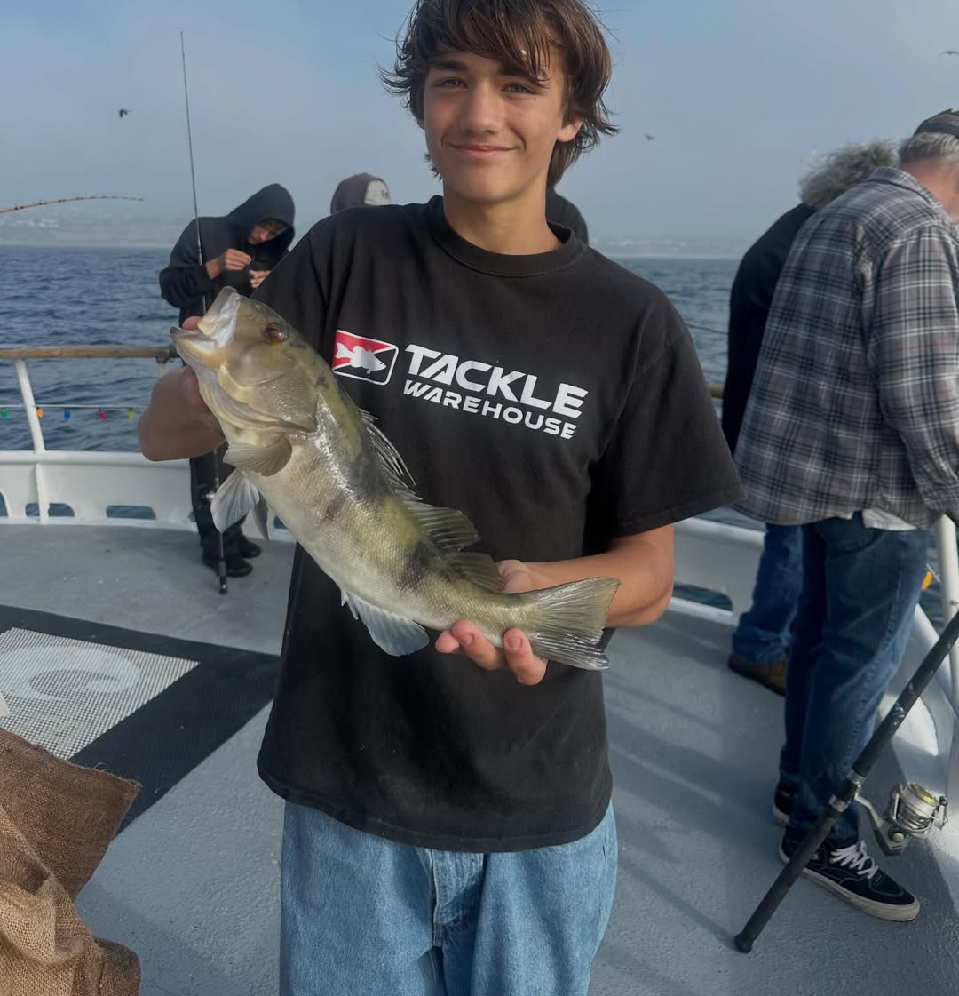 Person on a boat holding a fish, wearing a black T-shirt and jeans, with water in the background.