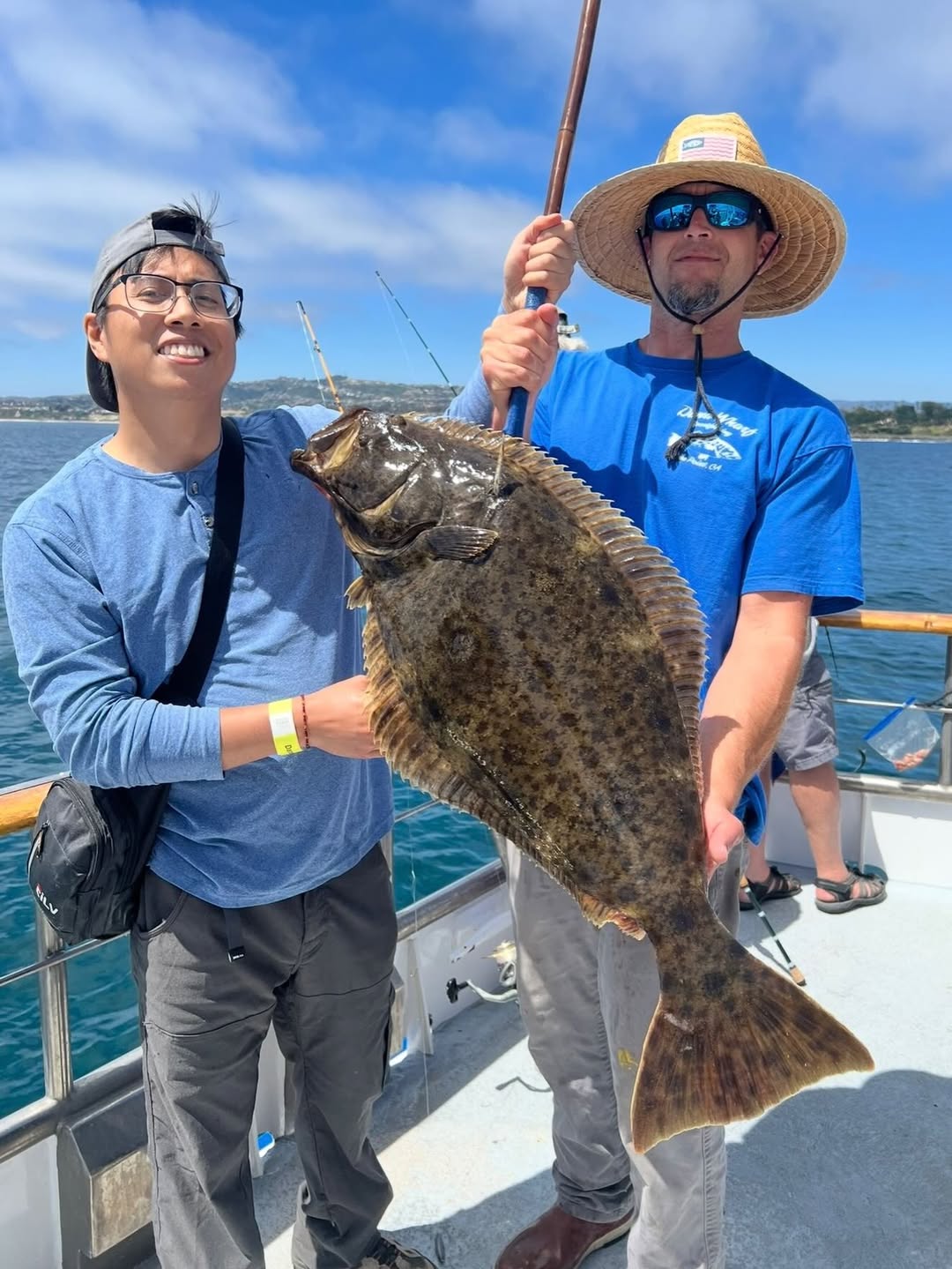 Two people on a boat holding a large flatfish, with blue sky and ocean in the background.