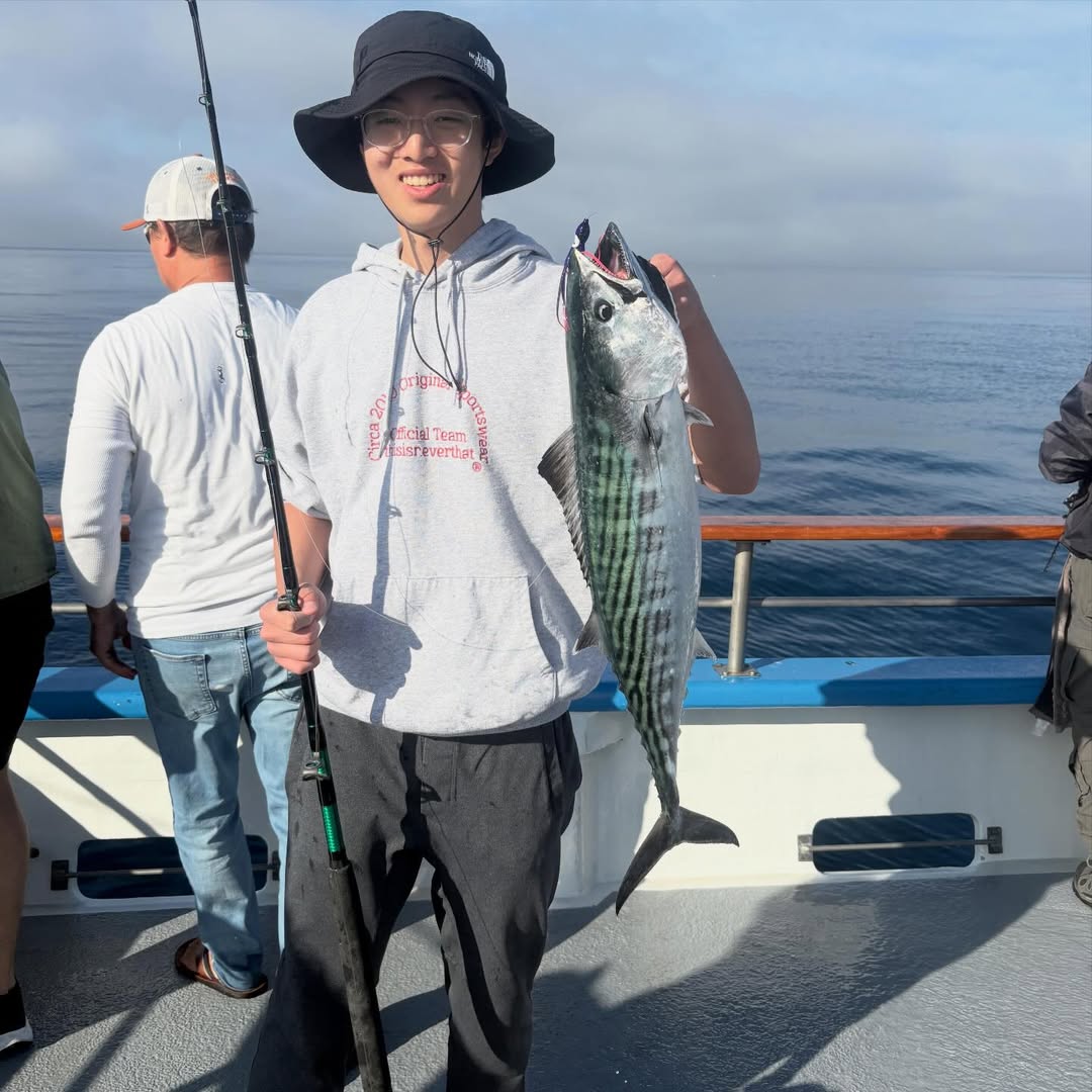 Person on a boat holding a large fish, wearing a hoodie and bucket hat, with water in the background.