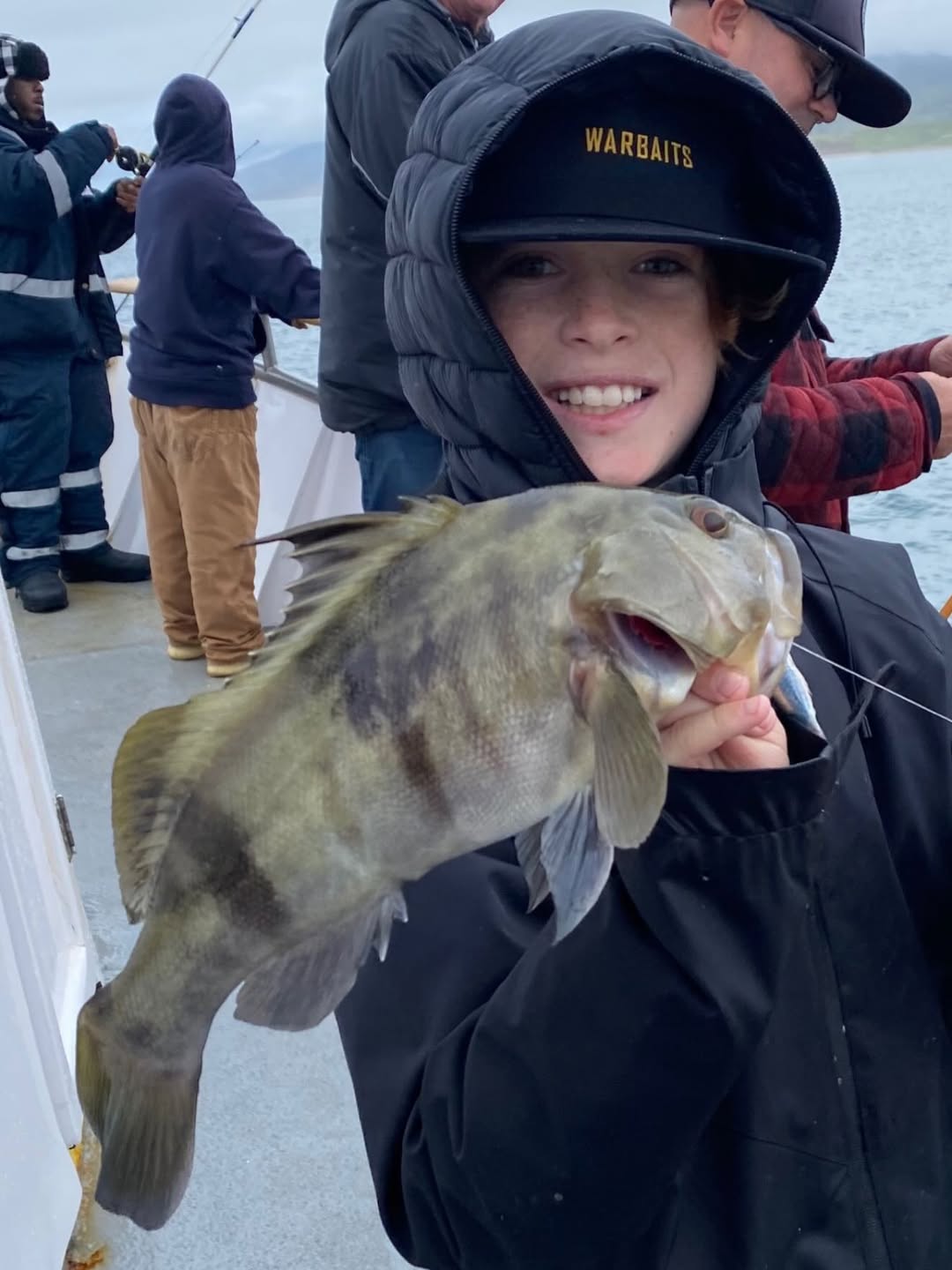 Person in hoodie and cap smiling, holding a fish on a boat.