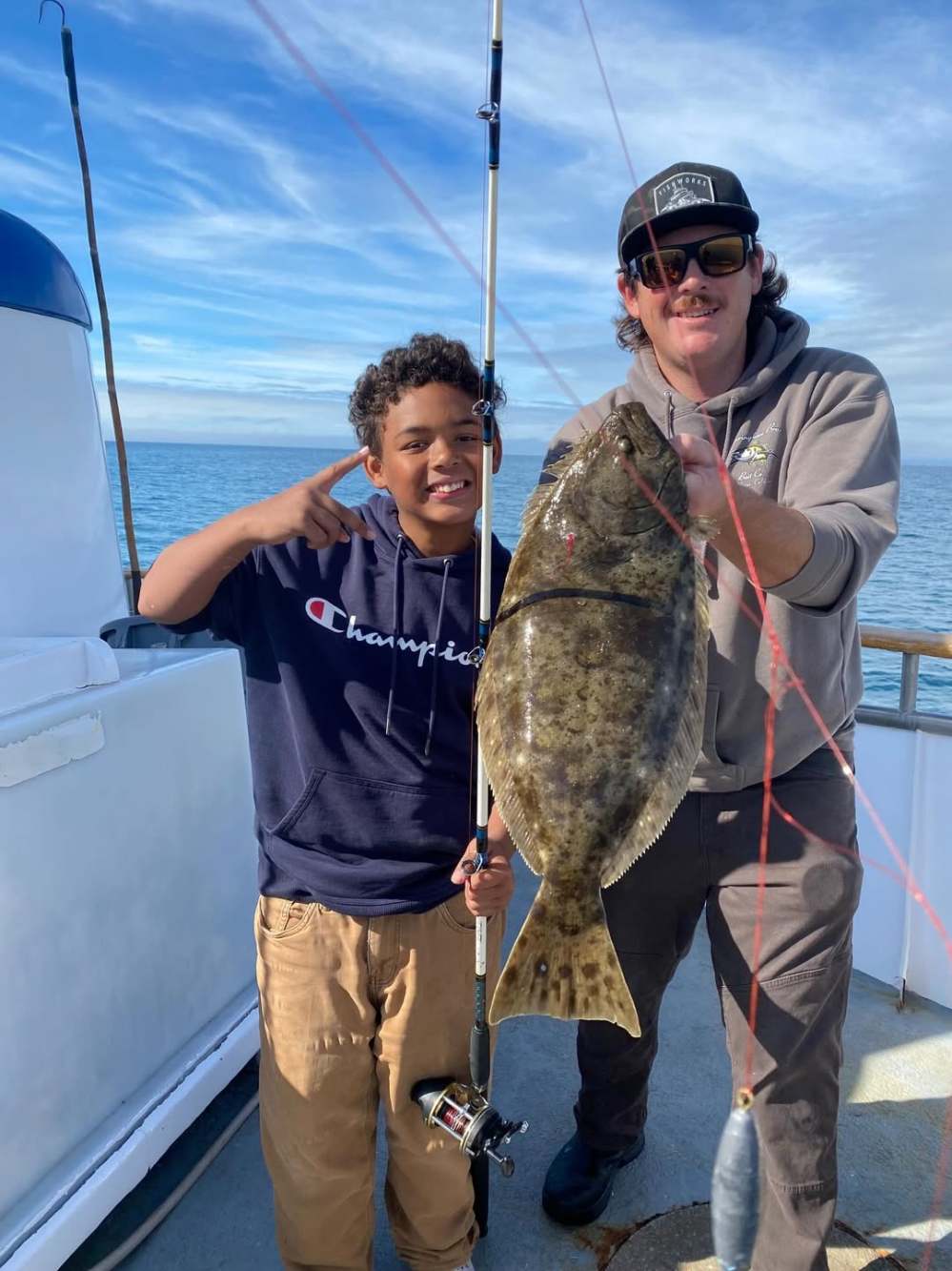 Two people smiling on a boat holding a large fish with a fishing rod, under a blue sky.