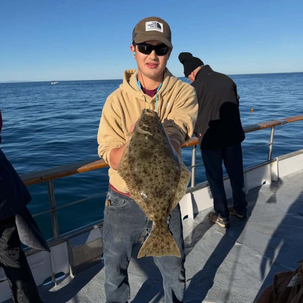 Person holding a large fish on a boat with two other people and the ocean in the background.
