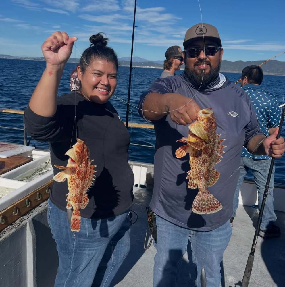 Two people on a boat holding colorful fish against a blue sky and ocean backdrop.