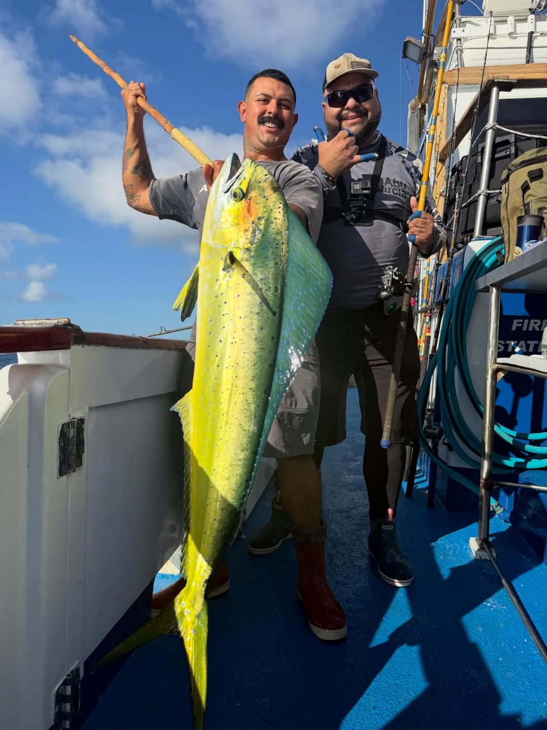 Two men on a boat, one holding a large green and yellow fish against a blue sky backdrop.