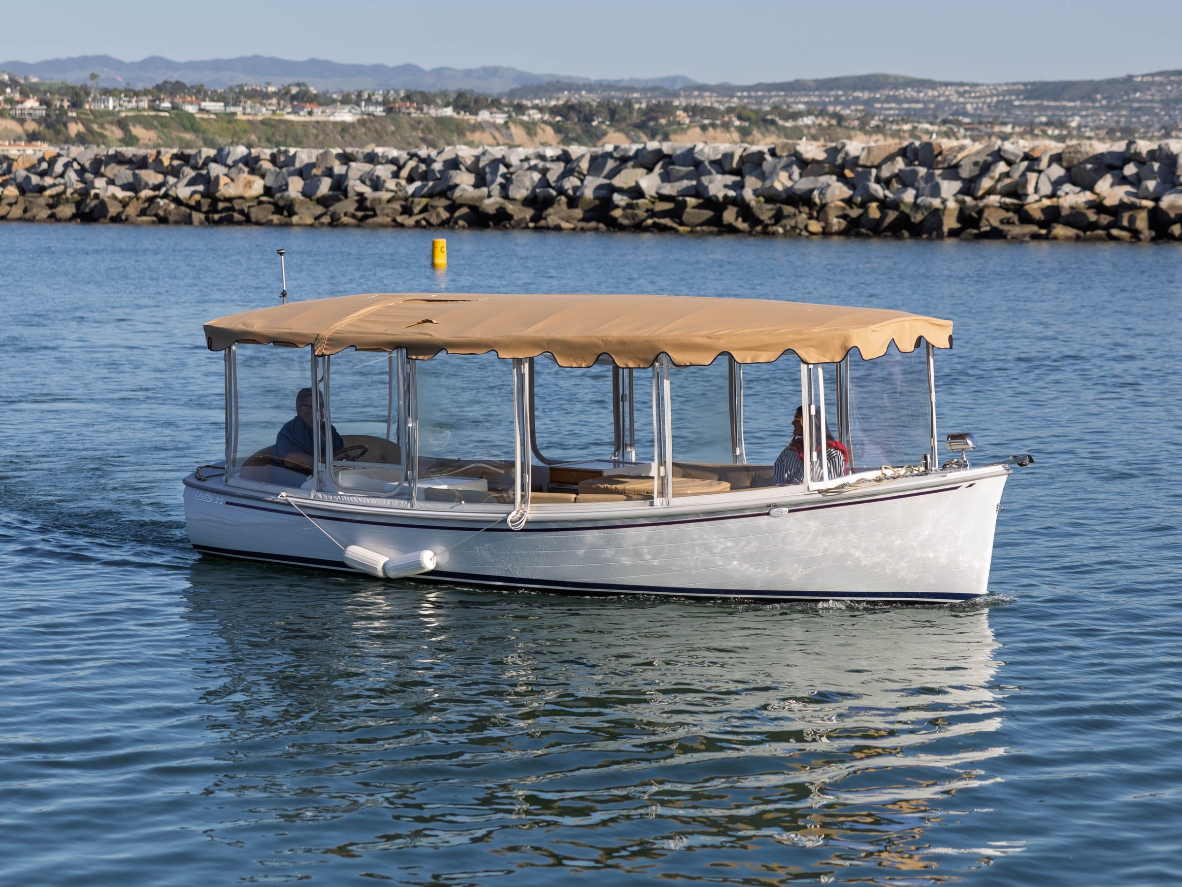 A white boat with beige canopy cruising on calm water near rocky shoreline.