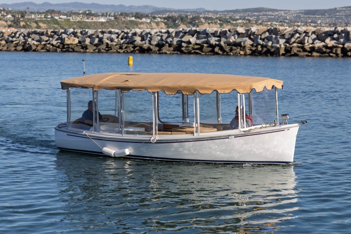 A white boat with beige canopy cruising on calm water near rocky shoreline.
