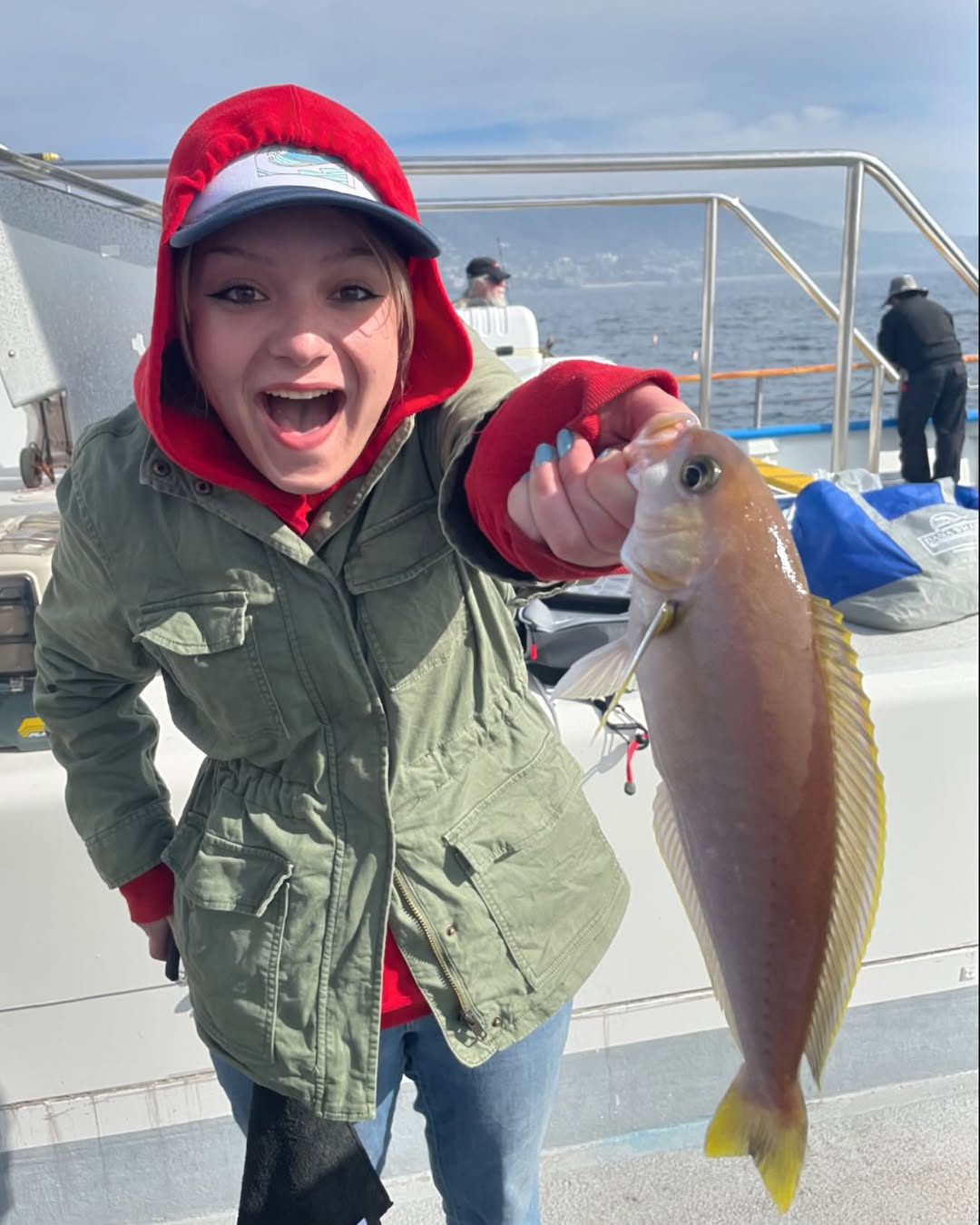 Person on a boat holding a fish, wearing a red hooded jacket and a cap, smiling excitedly at the camera.