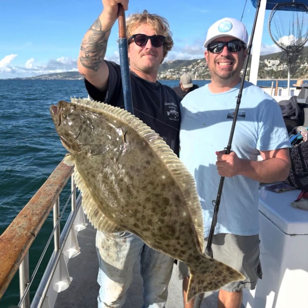Two men on a boat holding a large fish they caught, smiling at the camera.