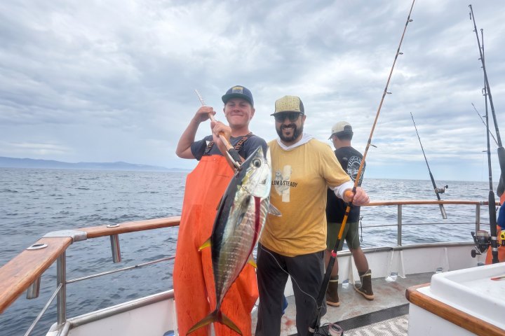Two people on a boat smiling, holding a large fish, cloudy sky above.